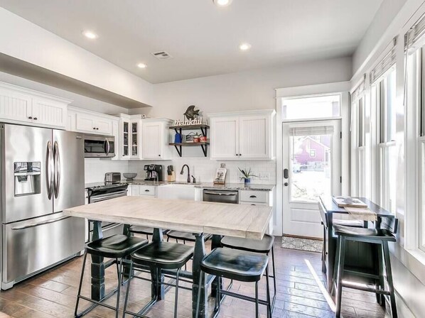 A modern kitchen with white cabinetry, stainless steel appliances, a large island with bar stools, and a view of the outside through large windows.