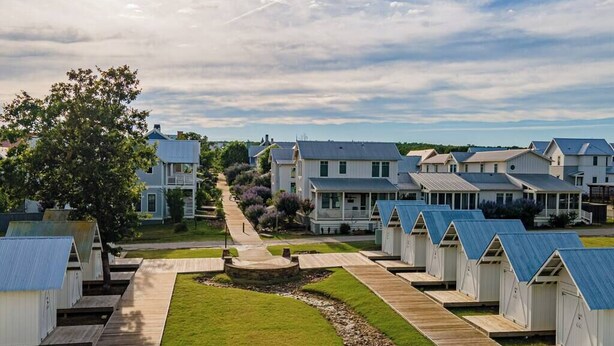 A row of beach houses with a wooden walkway leading to a central area, surrounded by greenery and a clear sky.