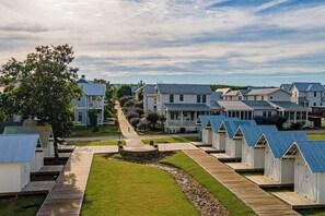 A row of beach houses with a wooden walkway leading to a central area, surrounded by greenery and a clear sky.
