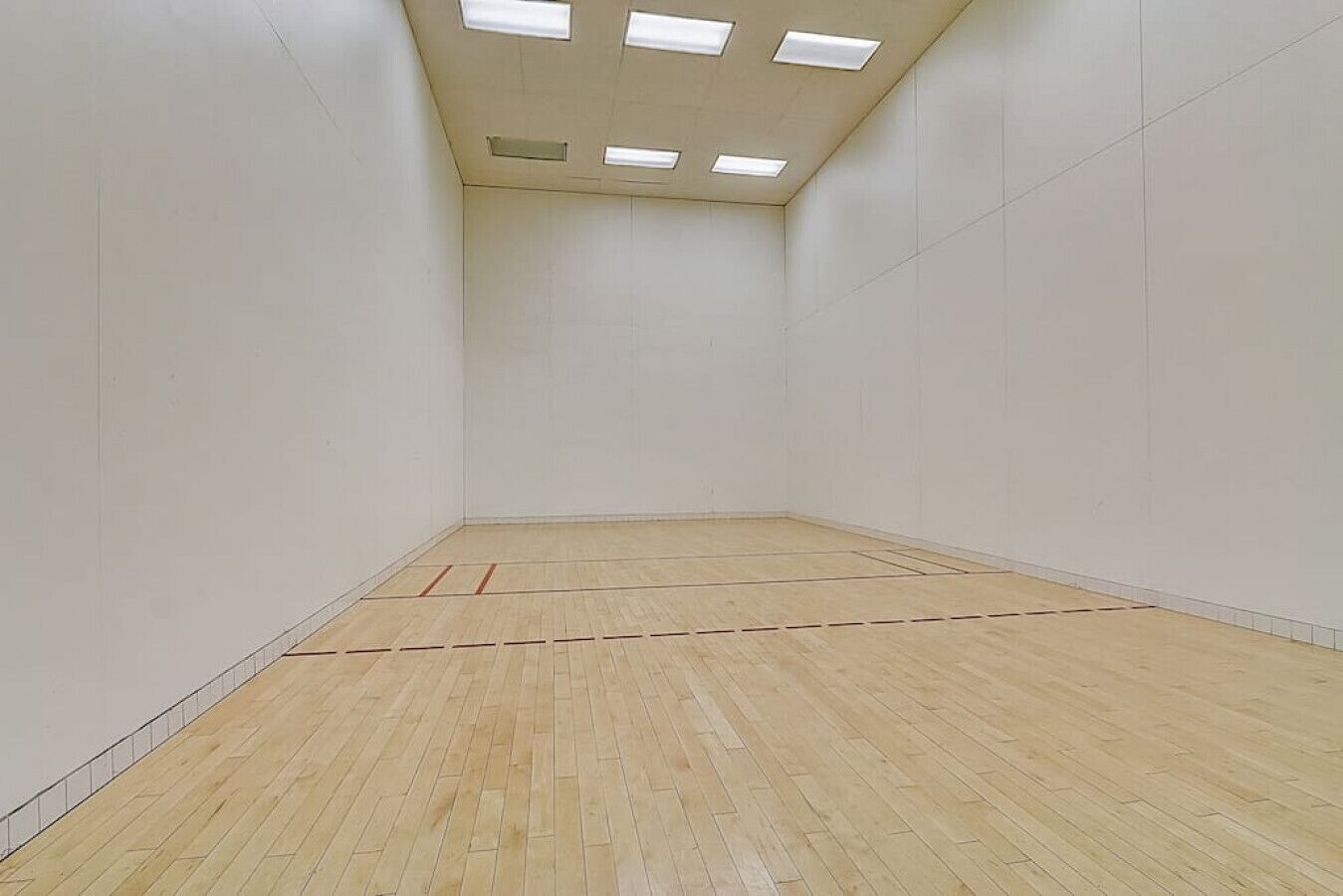 A squash court with a wooden floor, white walls, and a ceiling with rectangular lights.