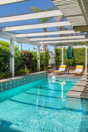 Una piscina azul cristalina con borde de azulejos, rodeada por una zona cubierta con dos sillas de descanso y una sombrilla verde.