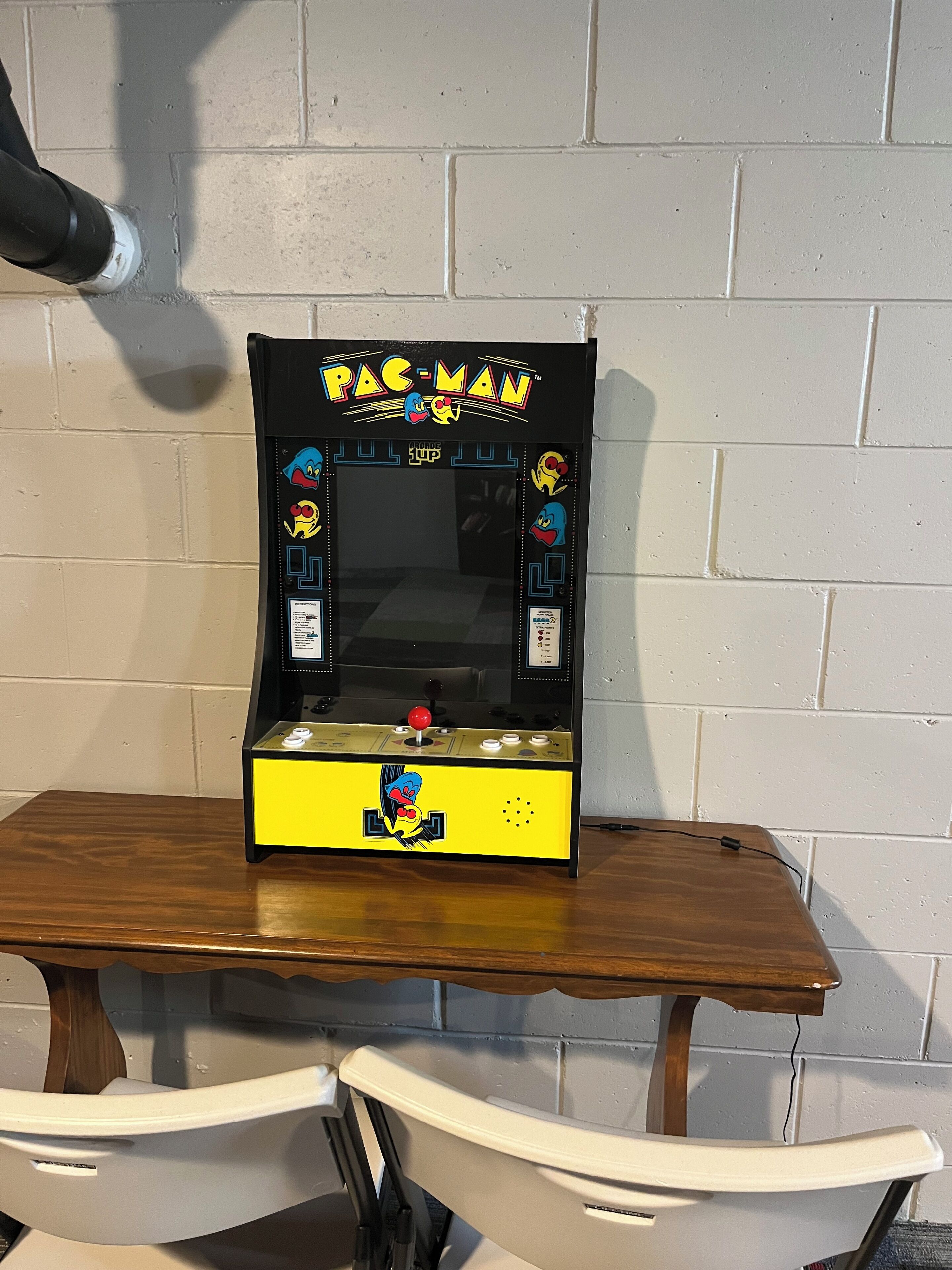 Une borne d’arcade Pac-Man posée sur une table en bois, adossée à un mur en briques blanches.