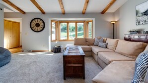 Ein Wohnzimmer mit Holzboden, einer großen Uhr, einem beigefarbenen Sofa, einem Couchtisch und Blick auf eine grüne Landschaft durch ein Erkerfenster.
