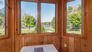 Ein Zimmer mit Holzvertäfelung, drei großen Fenstern, einem weißen Tisch und Blick auf eine grüne Wiese mit Bäumen.