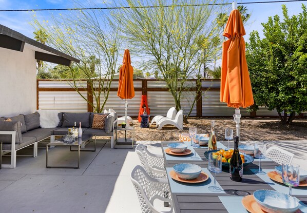 An outdoor patio with a dining table set for six, surrounded by white chairs. There are orange umbrellas, a sofa, and a small table with a bottle and glasses.