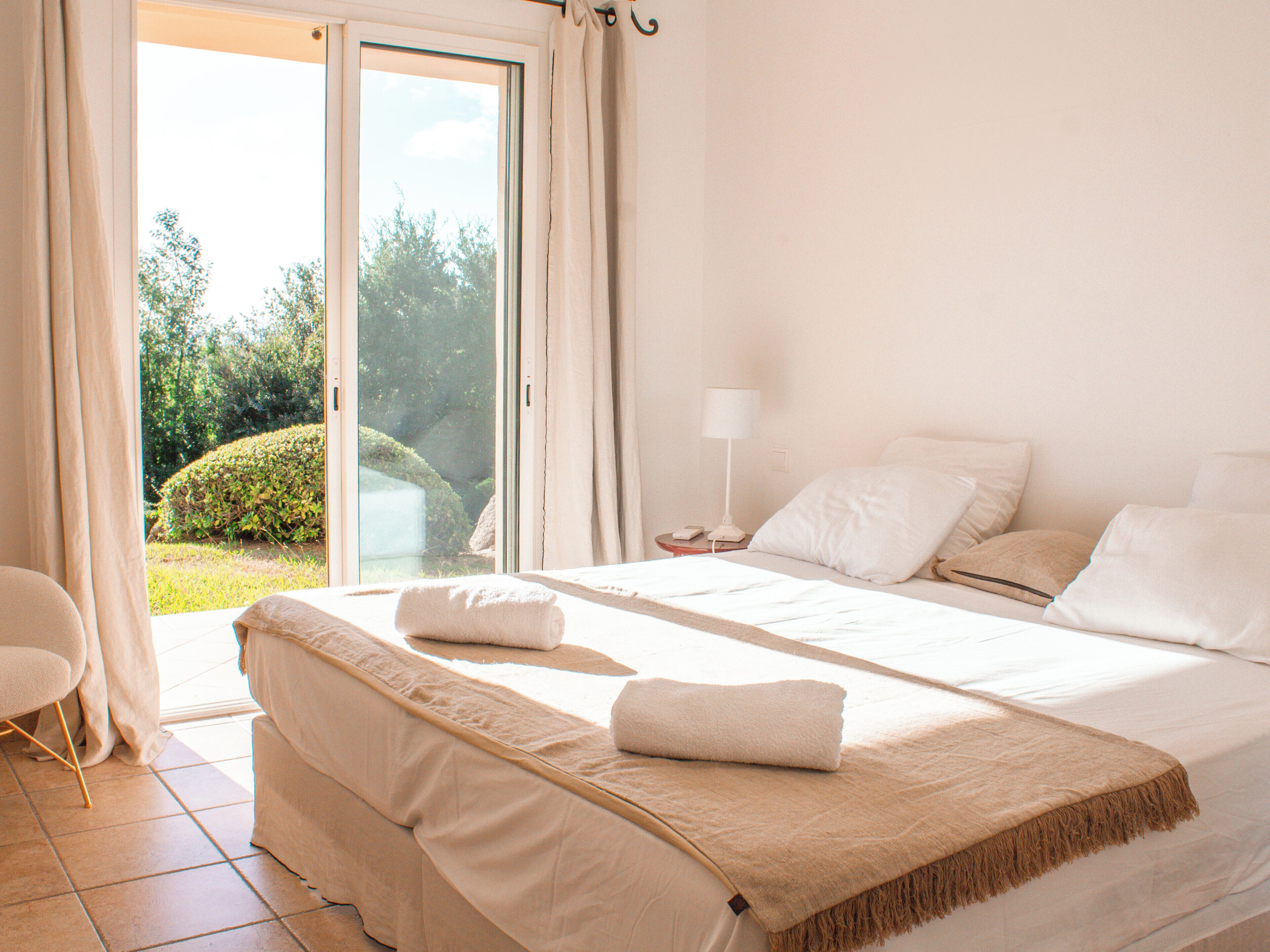 A bedroom with a bed, a nightstand, a lamp, and a view of the outdoors.