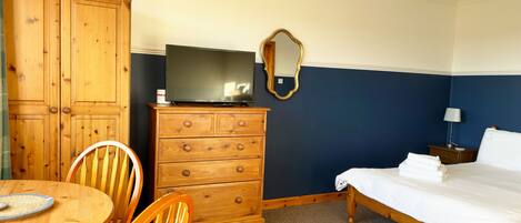 A hotel room with a wooden dresser, a television on top, a bed with white linens, a round wooden table with chairs, and a mirror on the wall.