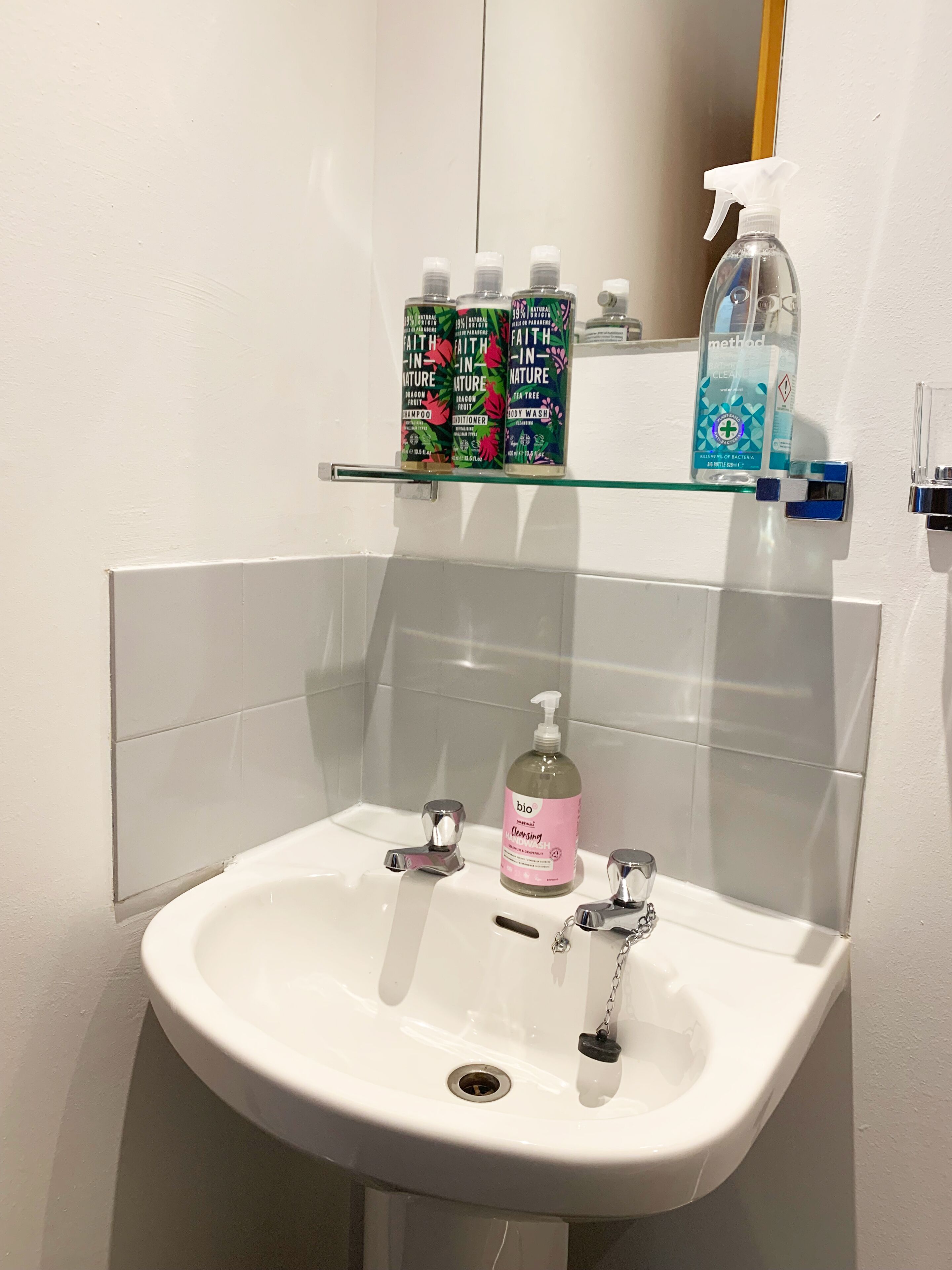 Un baño con lavamanos blanco, dos llaves y un estante de vidrio arriba con varios artículos de tocador.
