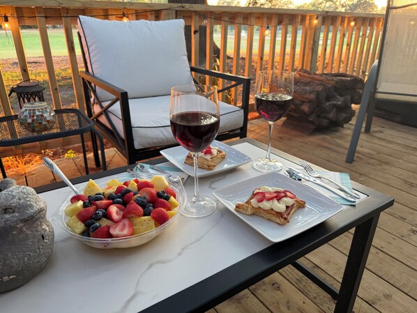 A table with fruit salad, two glasses of red wine, and a pastry on a wooden deck with a chair and a lamp in the background.