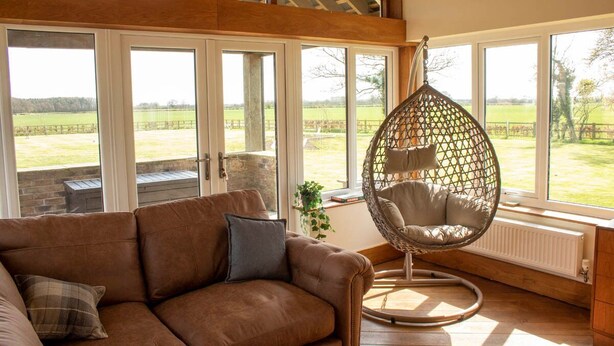 A cozy room with a brown leather sofa, a hanging chair, and large windows offering a view of a green field. - Luxury Farmhouse:Padel court/Hot tub/pool table/fire pit. Central meet up point. (SELBY)