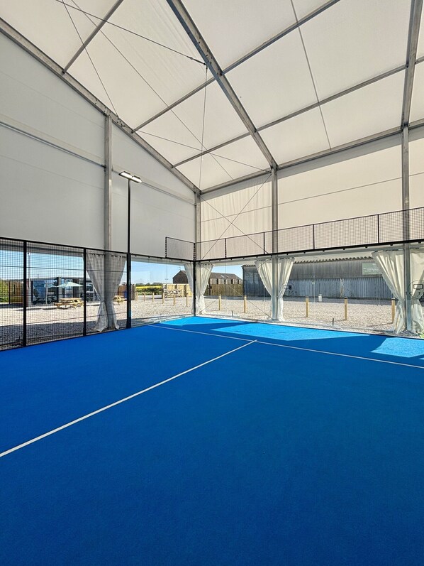 An indoor tennis court with a blue surface, enclosed by a netted fence and a white roof structure.
