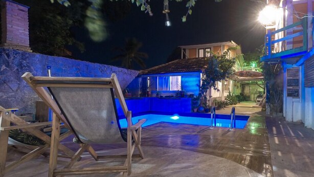 Una zona de piscina con una silla de madera, una casa con iluminación azul y un muro de piedra.