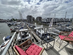 A marina with boats docked, a wooden deck with wicker chairs and a table, and buildings in the background.