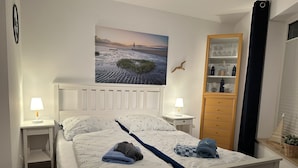 Ein ordentlich bezogenes Bett mit einem maritime-inspirierten Bettbezug, zwei Nachttischlampen, ein Holznachttisch, eine Holzkommode und ein gerahmtes Bild einer Küstenlandschaft an der Wand.