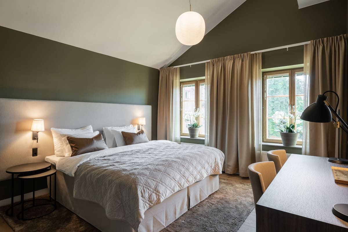 Une chambre à coucher comprenant un lit, une table de chevet, une lampe, un bureau, une chaise et une vue sur la verdure par la fenêtre.