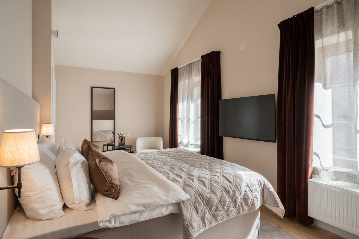 Une chambre d’hôtel avec un lit, une télévision, une lampe, un miroir et une fenêtre avec des rideaux.