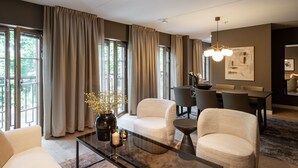 A modern living room with a glass coffee table, beige armchairs, a dining area with a dark table, and large windows with curtains. - Live and Stay - Ekmanska (Gothenburg)