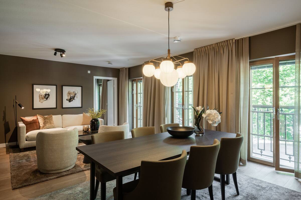 Un espace de salle à manger moderne, doté d’une grande table, de chaises et d’un lustre. On y trouve également un canapé et une table basse agrémentée d’objets décoratifs.