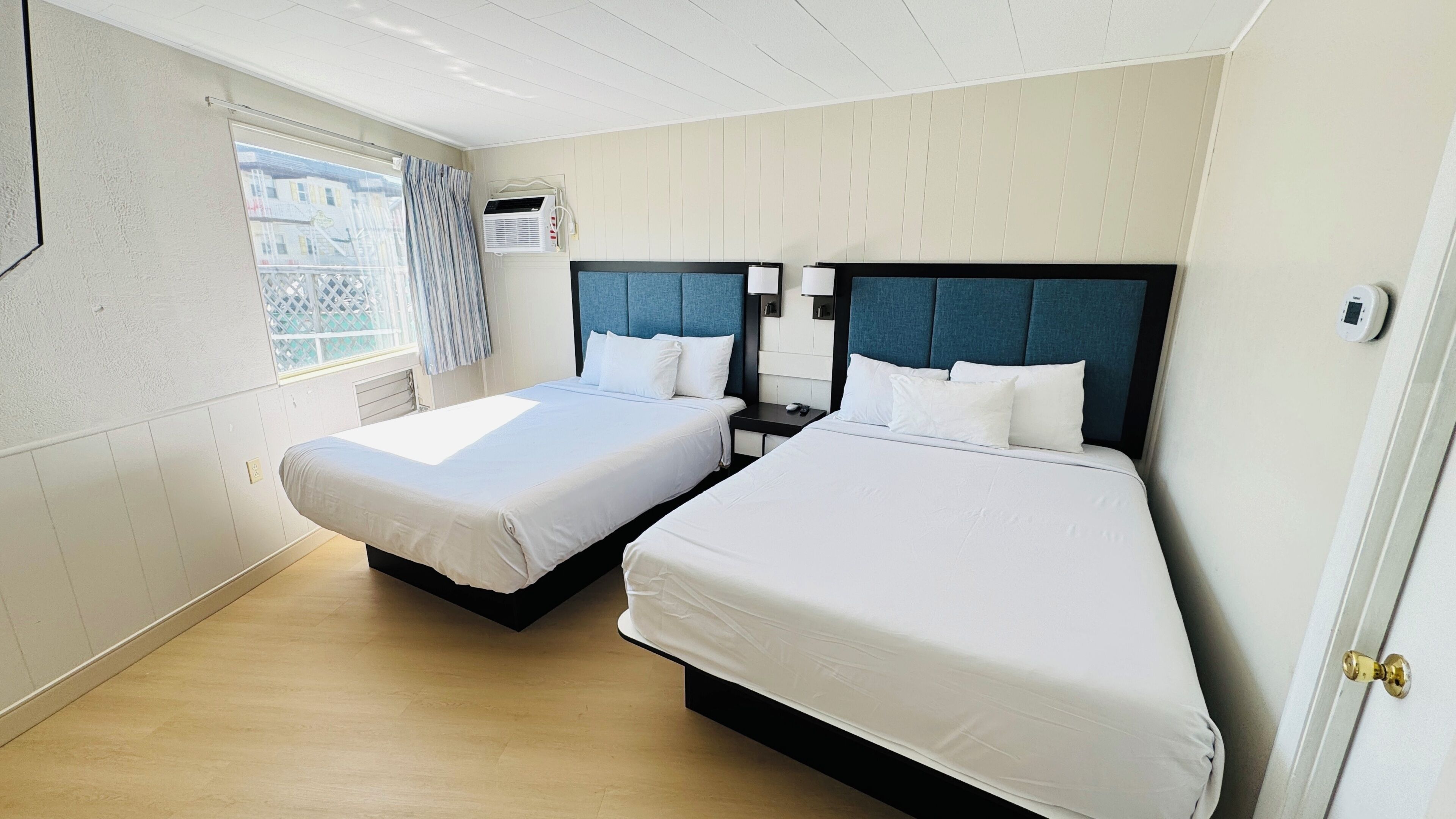 A hotel room with two beds, a window with curtains, an air conditioning unit, and a bedside table with a lamp.