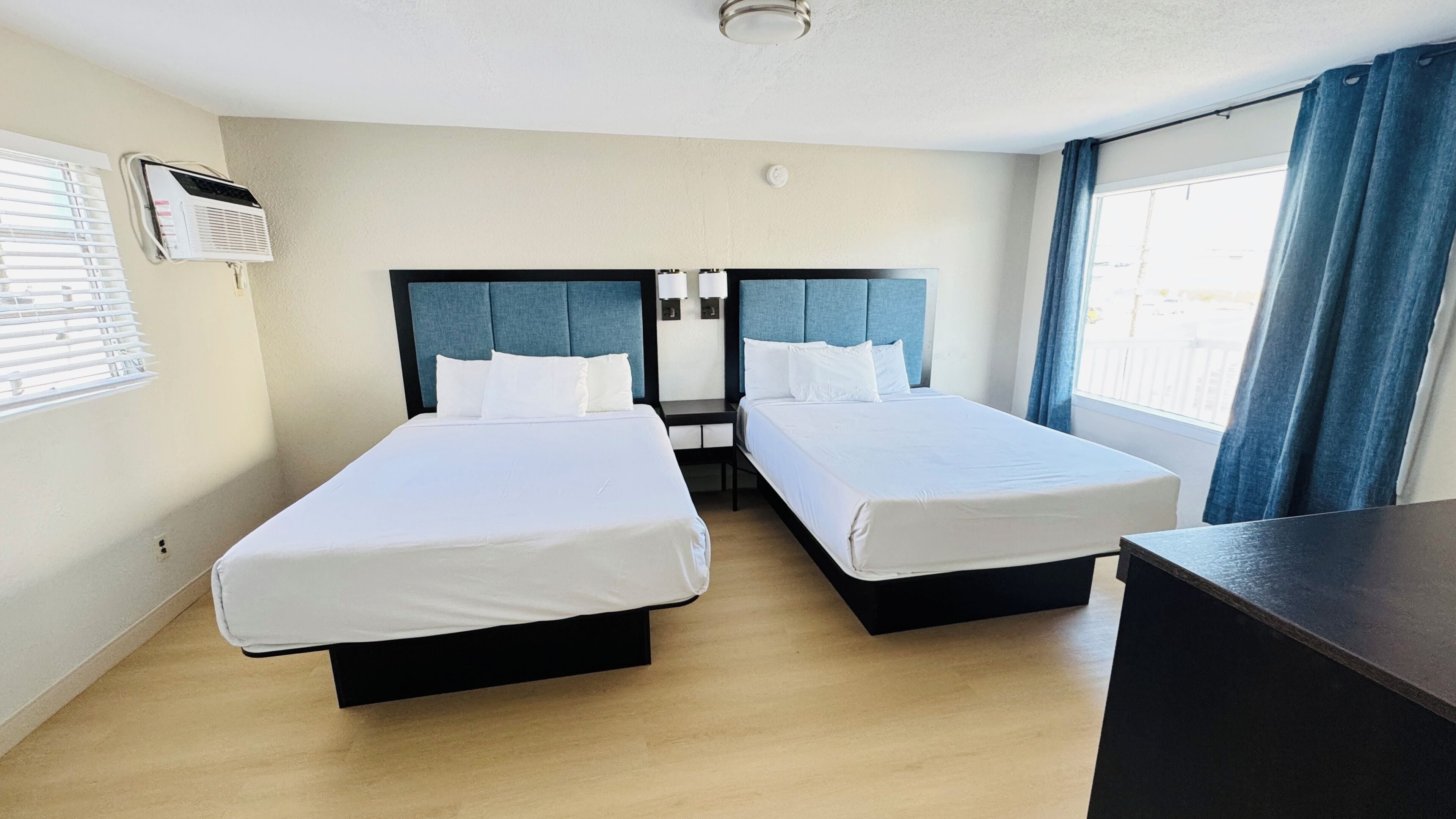 A hotel room with two beds, a desk, a window with curtains, and an air conditioning unit.