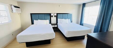 A hotel room with two beds, a desk, a window with curtains, and an air conditioning unit.