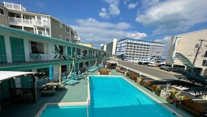 A pool area with lounge chairs and umbrellas, a building with balconies, and a street with parked cars. - Bel Air Motel (Wildwood Crest)