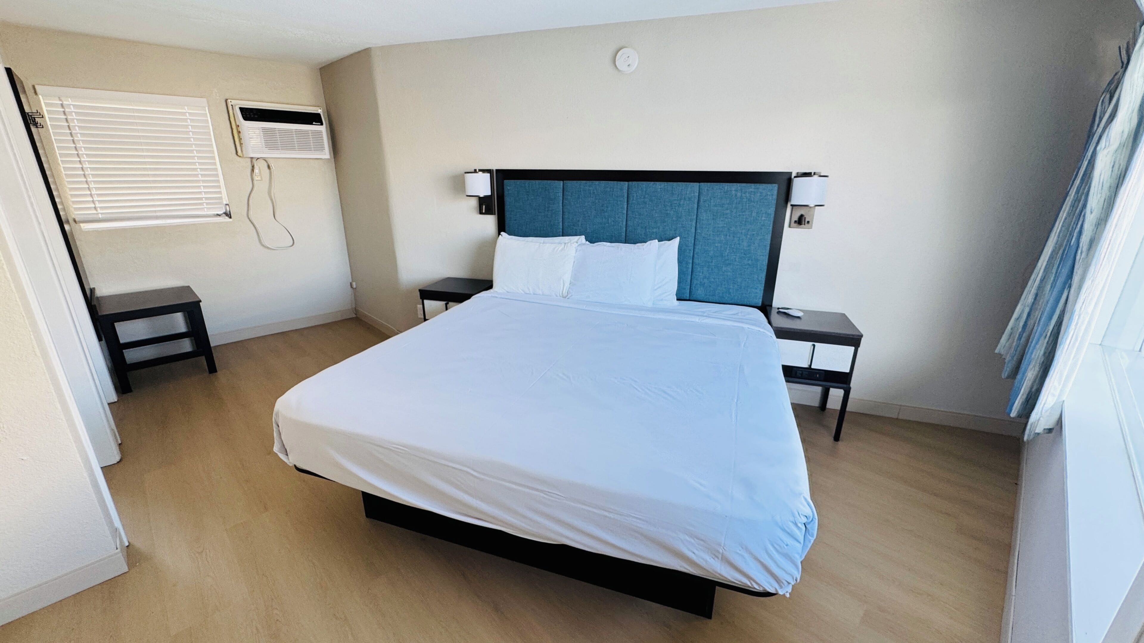 A neatly arranged hotel room with a bed, bedside tables, a small stool, a window with blinds, and an air conditioning unit.