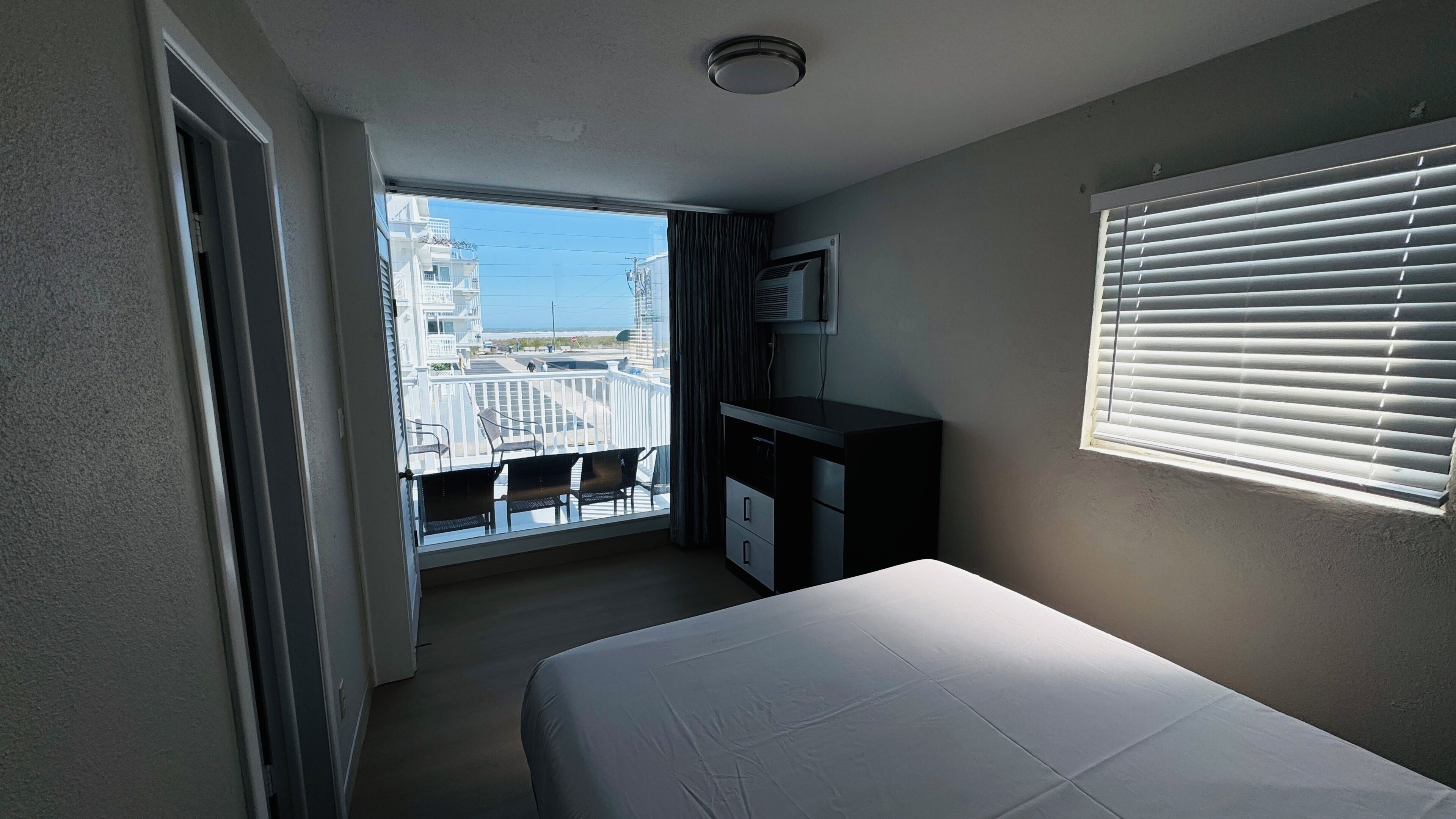 A hotel room with a bed, a desk, a window with blinds, and a view of buildings and a balcony.