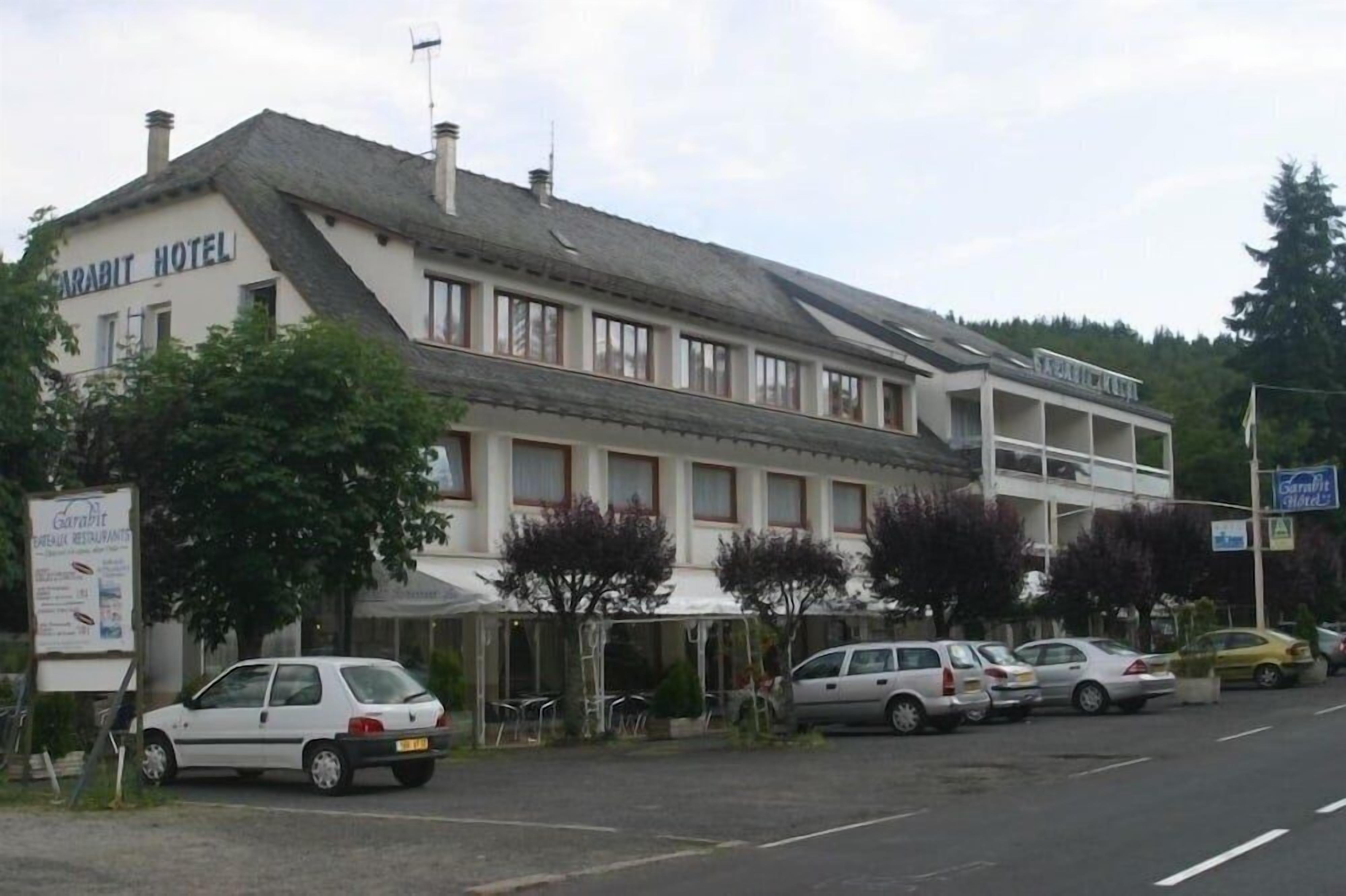 Hotel Garabit de dois andares, com fachada branca e telhado cinzento. Vários carros estão estacionados em frente, e uma placa anuncia o restaurante do hotel.