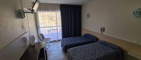 Habitación de hotel con dos camas, un escritorio con silla, una lámpara y un ventanal con cortinas.