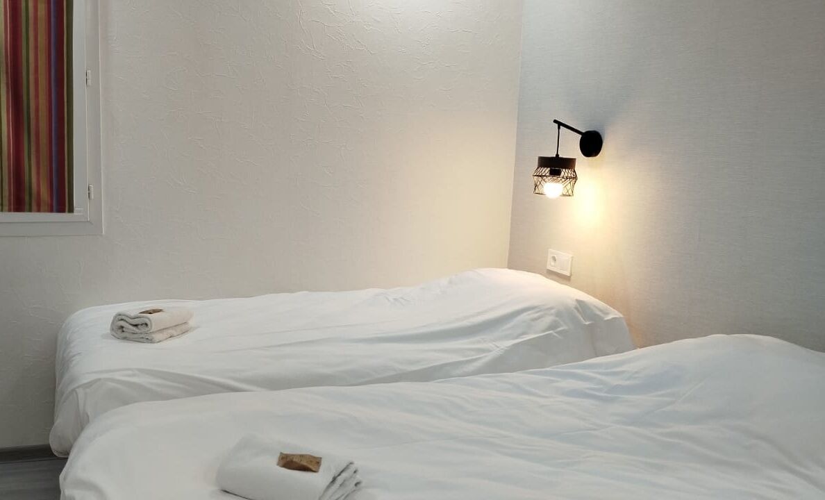 Une chambre d’hôtel avec deux lits simples, un luminaire fixé au mur et une fenêtre avec un rideau à rayures.