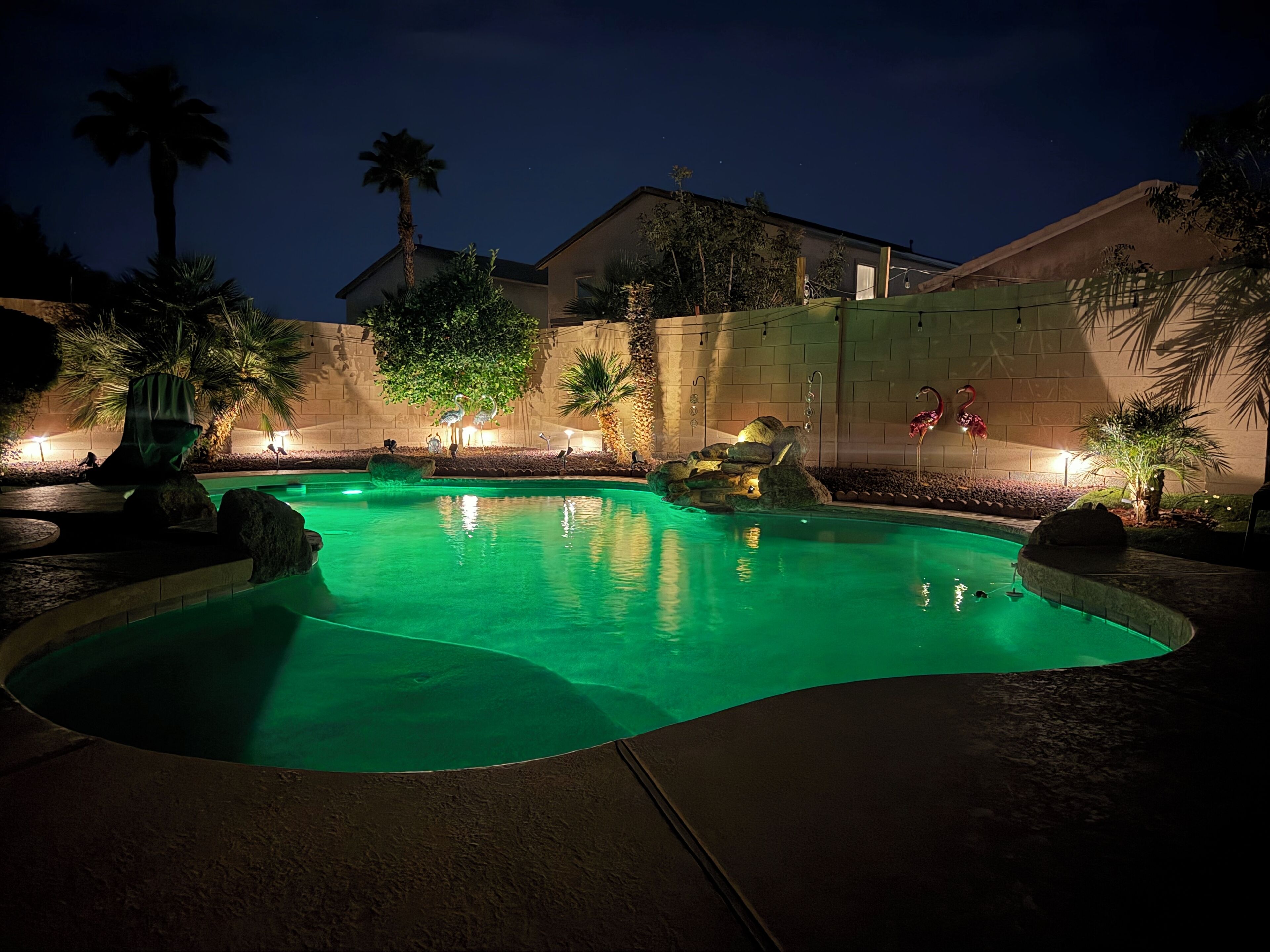 A nighttime swimming pool with illuminated rocks and a flamingo statue, surrounded by palm trees and a brick wall.