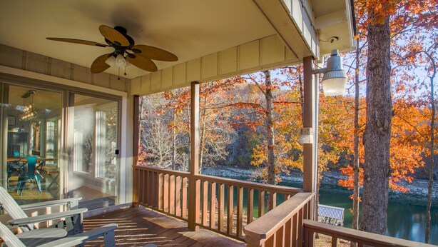 A wooden deck with Adirondack chairs, a ceiling fan, and a view of autumn trees and a lake.