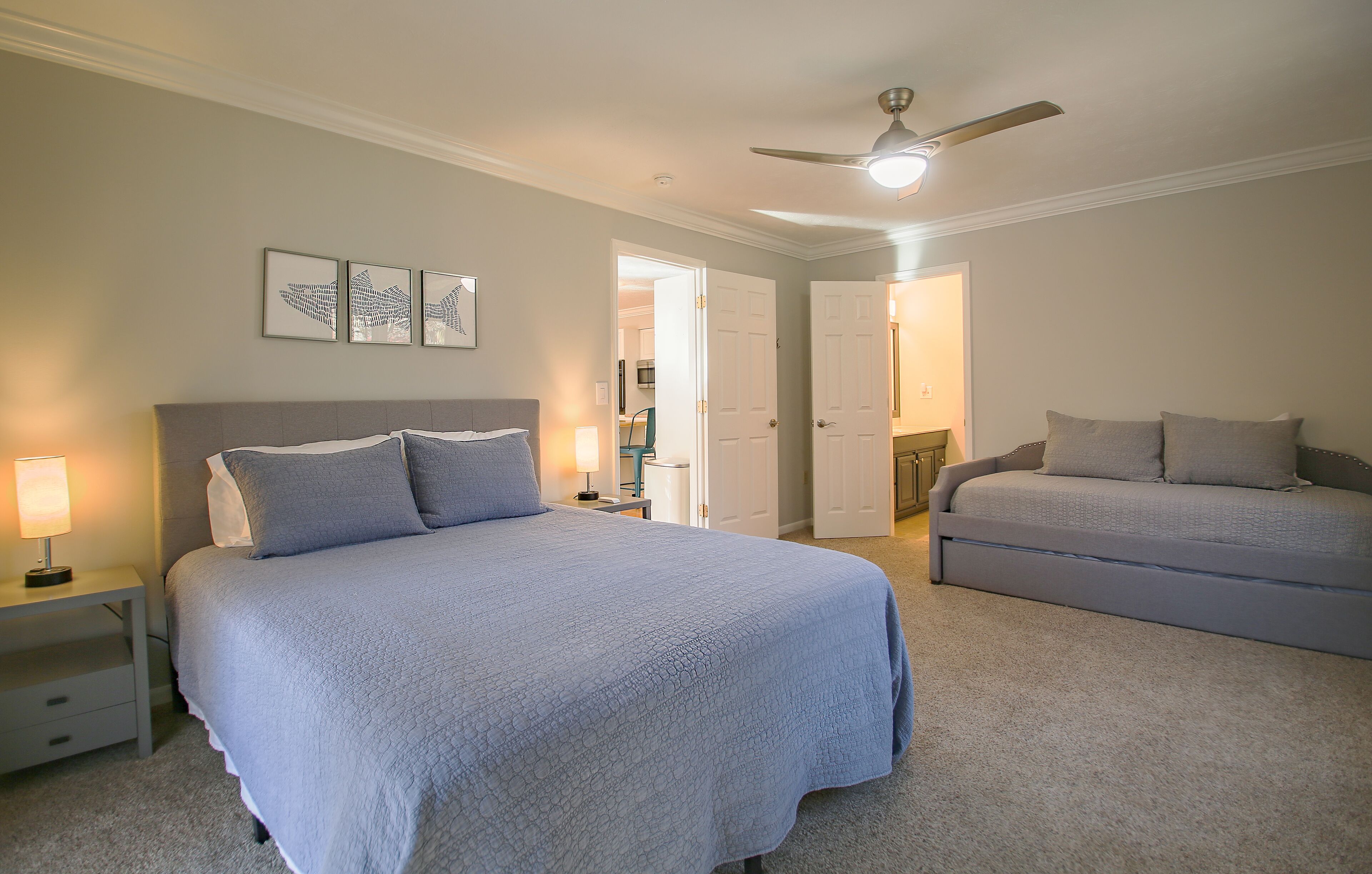 A bedroom with a large bed, a smaller sofa, a ceiling fan, and a door leading to another room.