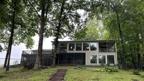 A modern two-story house with a large balcony, surrounded by trees and a grassy area with a wooden pathway leading to the entrance.