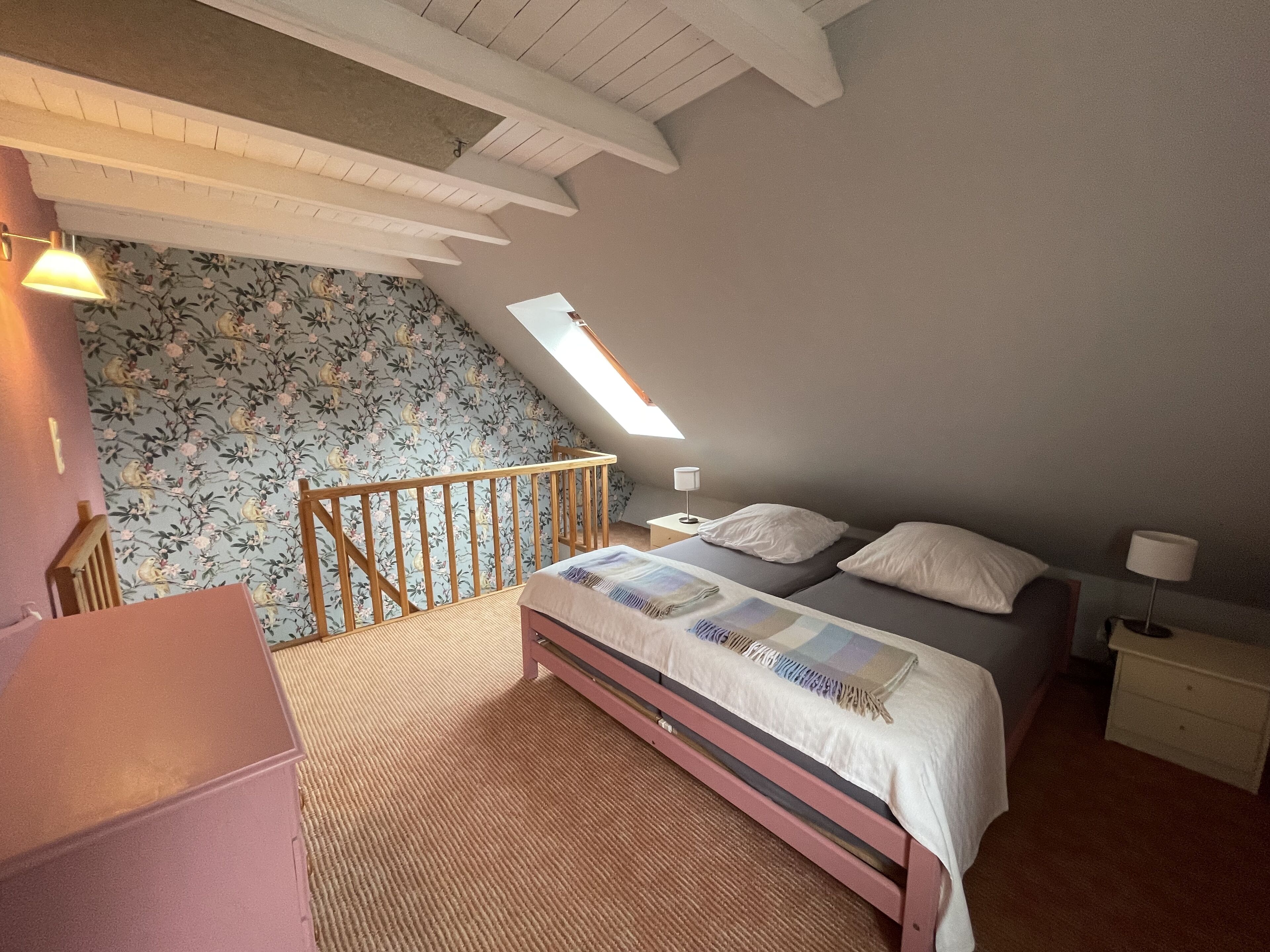 A bedroom with a sloped ceiling, a wooden headboard, a bed with a white and blue patterned bedspread, a nightstand with a lamp, and a small dresser.