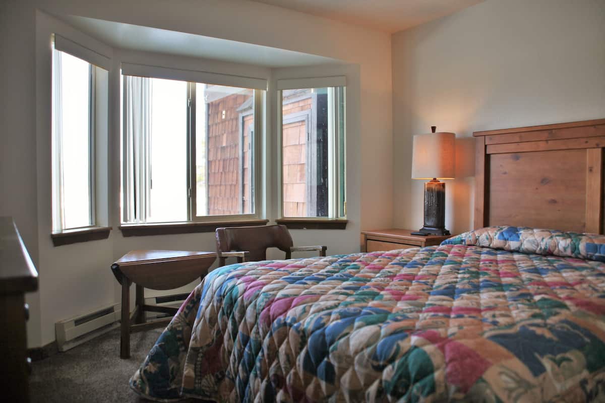 Une chambre avec un grand lit, un bureau et deux chaises. Elle est dotée de trois fenêtres qui laissent entrer la lumière naturelle.