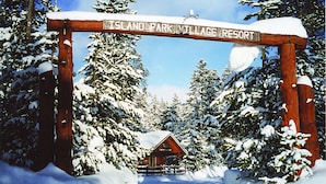 Um arco de madeira com a inscrição "Island Park Village Resort" que leva a uma cabana cercada por árvores cobertas de neve.