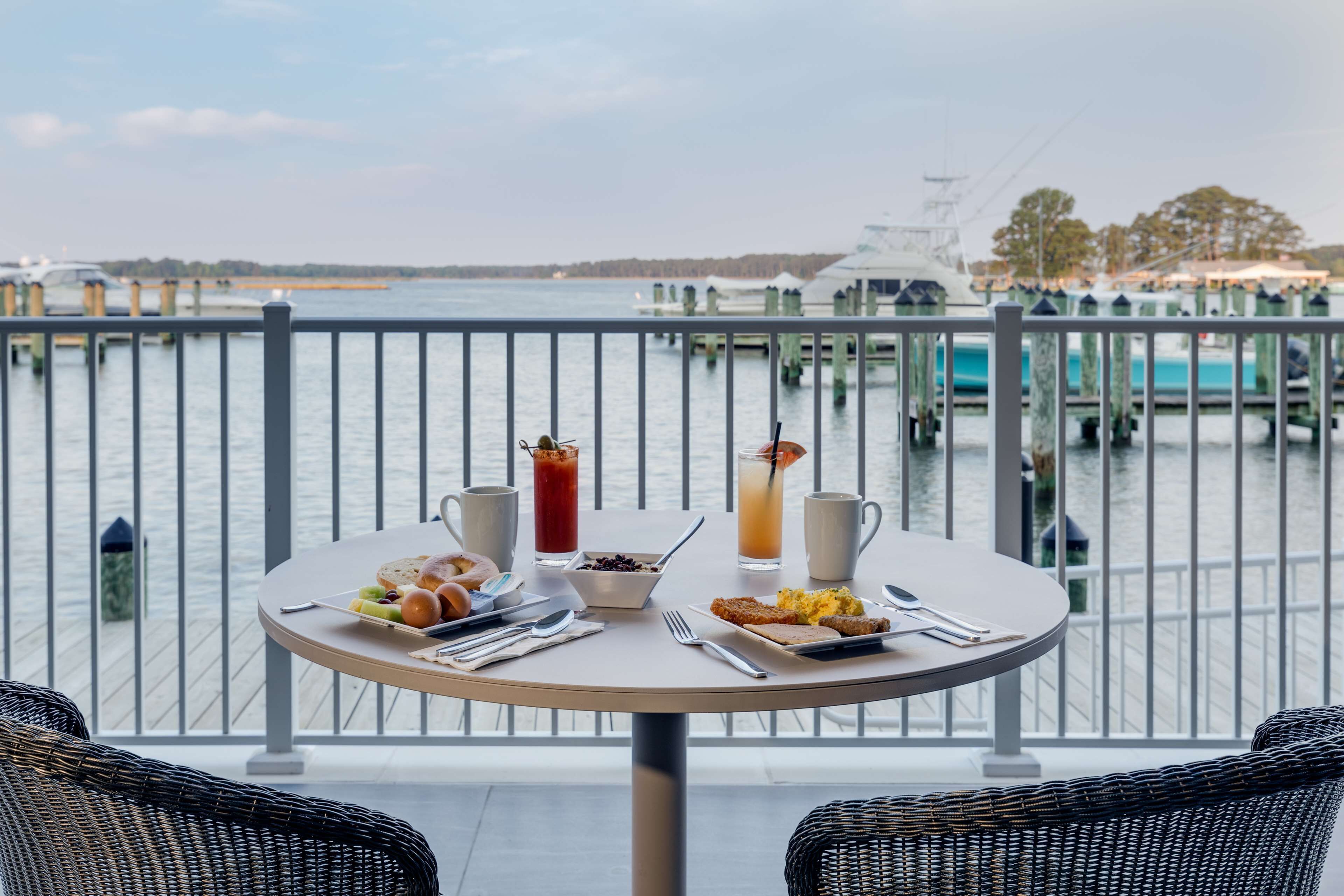 Mesa redonda con alimentos para el desayuno y bebidas, con vista a una marina con barcos y muelles.