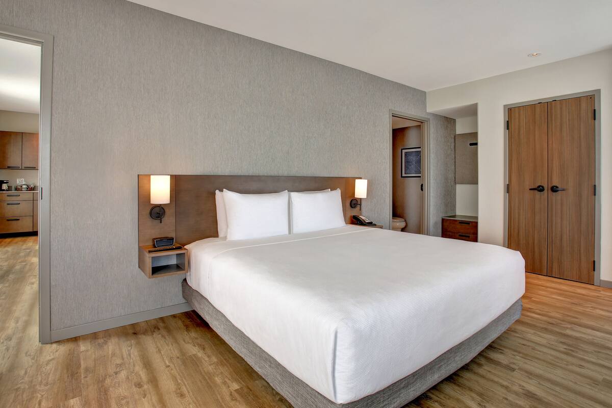 A modern hotel room with a large bed, a bedside table with a lamp, and a wooden wardrobe.