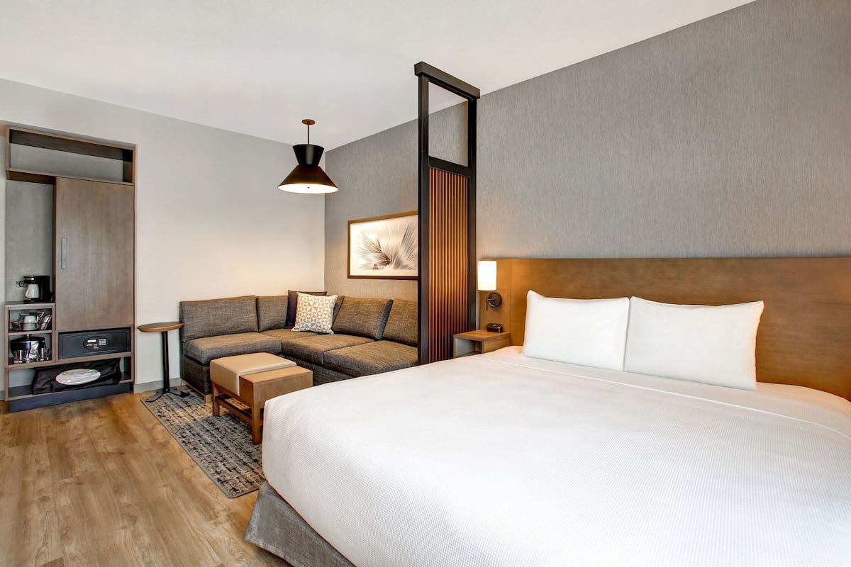 A modern hotel room with a large bed, a sofa, a small table, a nightstand, a mirror, and a wall-mounted lamp.