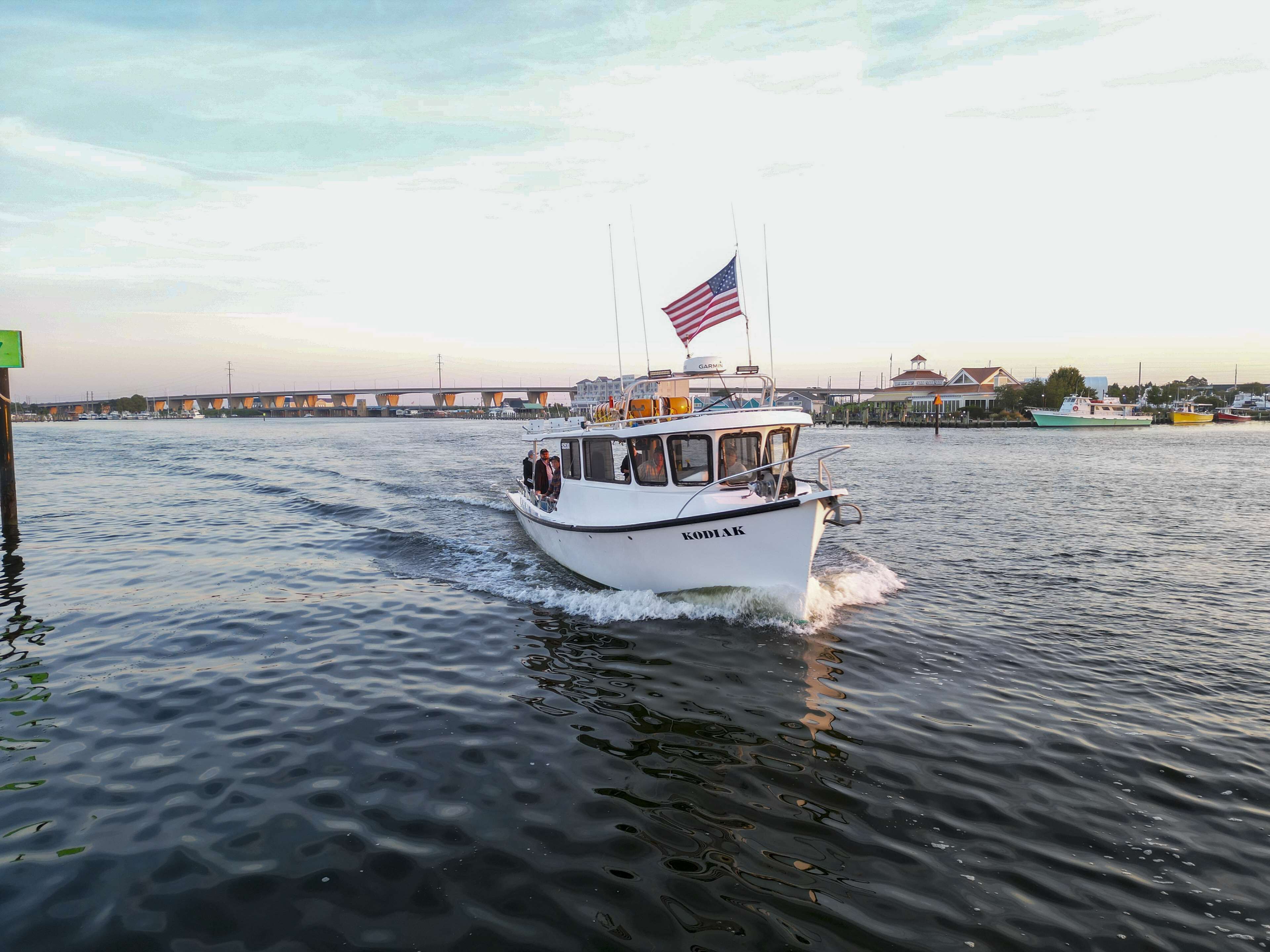 A boat named 'KODIAK' with an American flag, moving on a body of water with a bridge and buildings in the background.