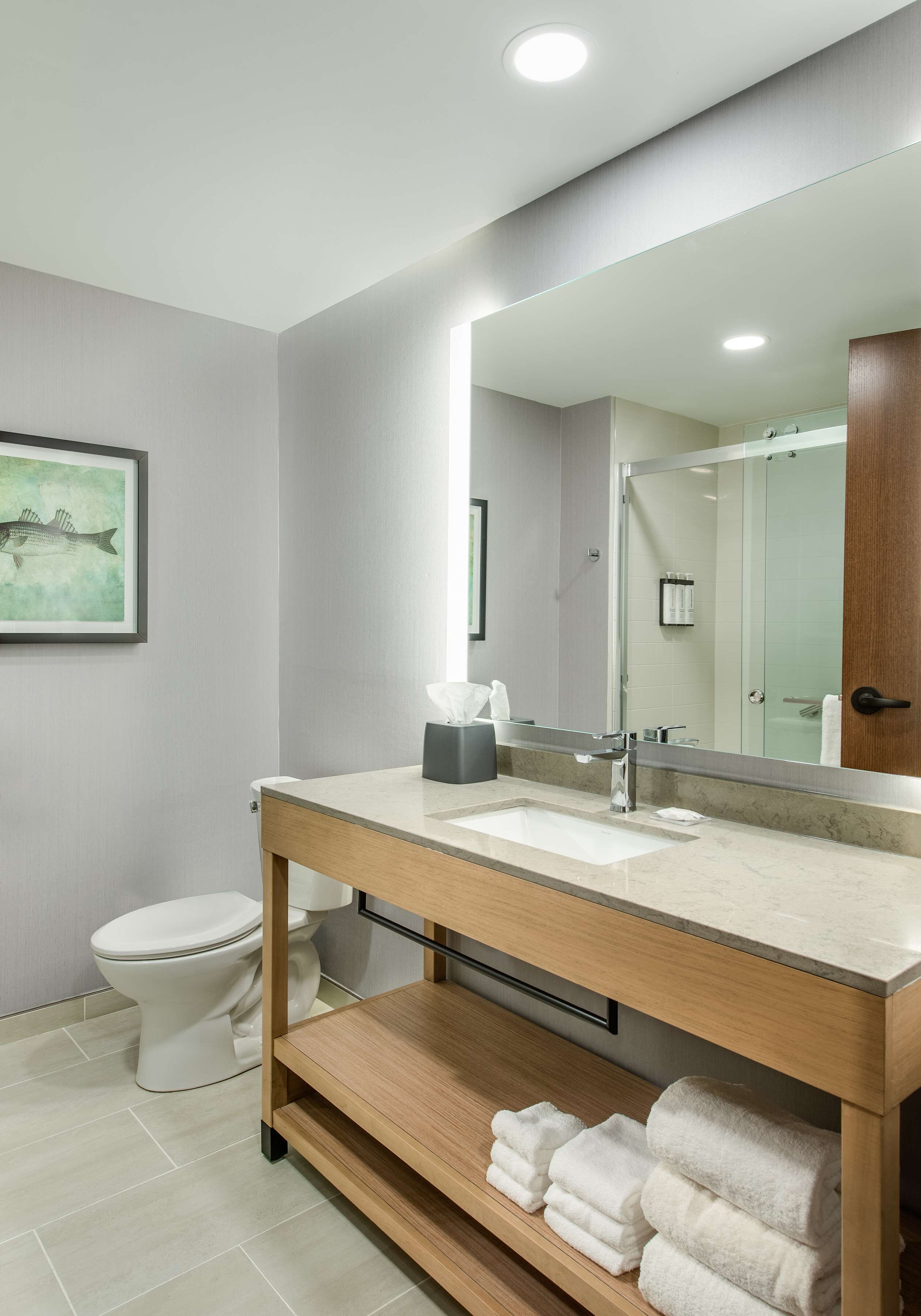 A modern bathroom with a large mirror, a sink, and a toilet. There is a towel rack with folded towels and a framed picture on the wall.