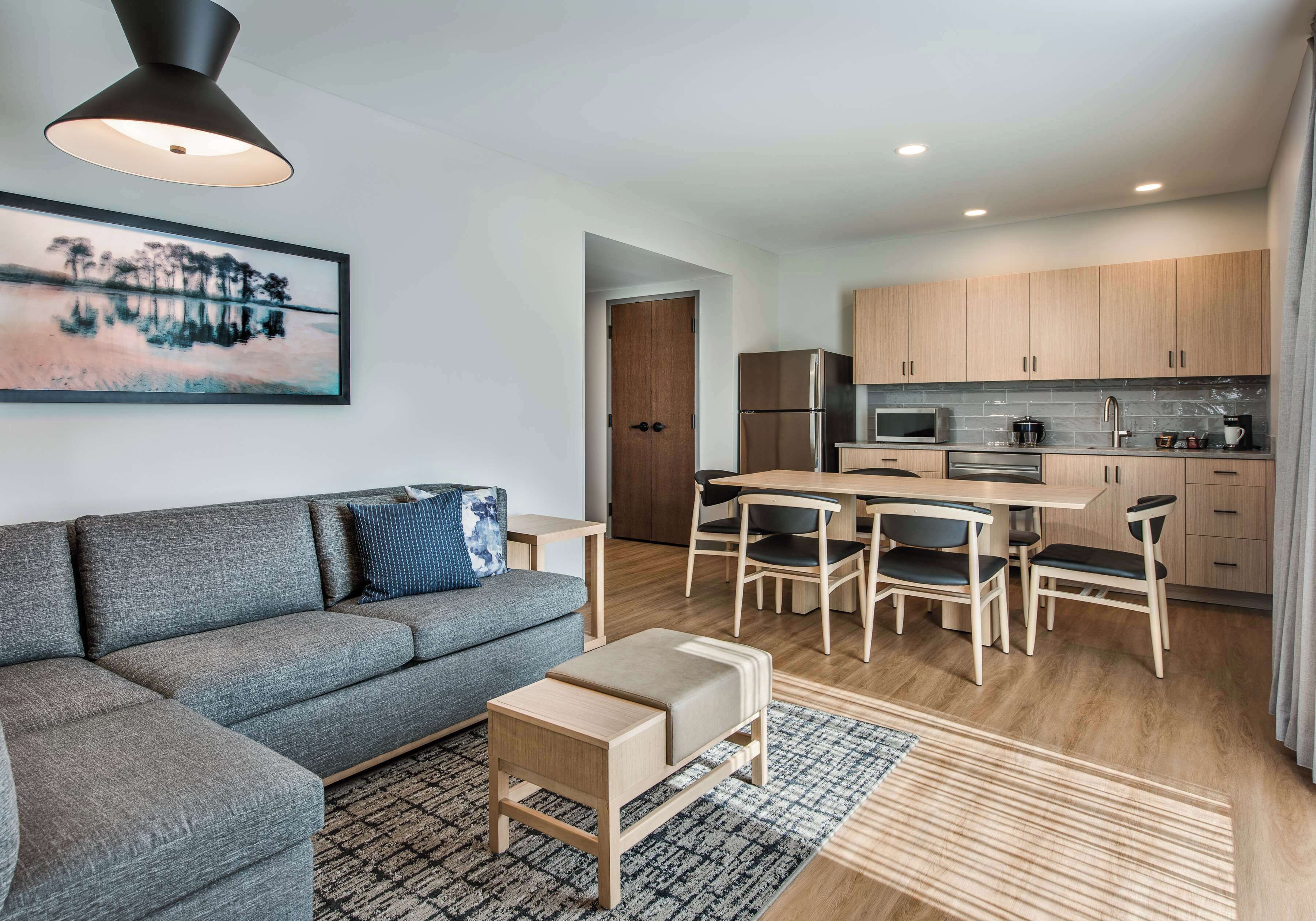 Una sala de estar moderna con un sofá gris, una mesa de centro de madera y un cuadro en la pared. Junto a ella, una cocina compacta con armarios de madera, un refrigerador y un comedor con una mesa y sillas.