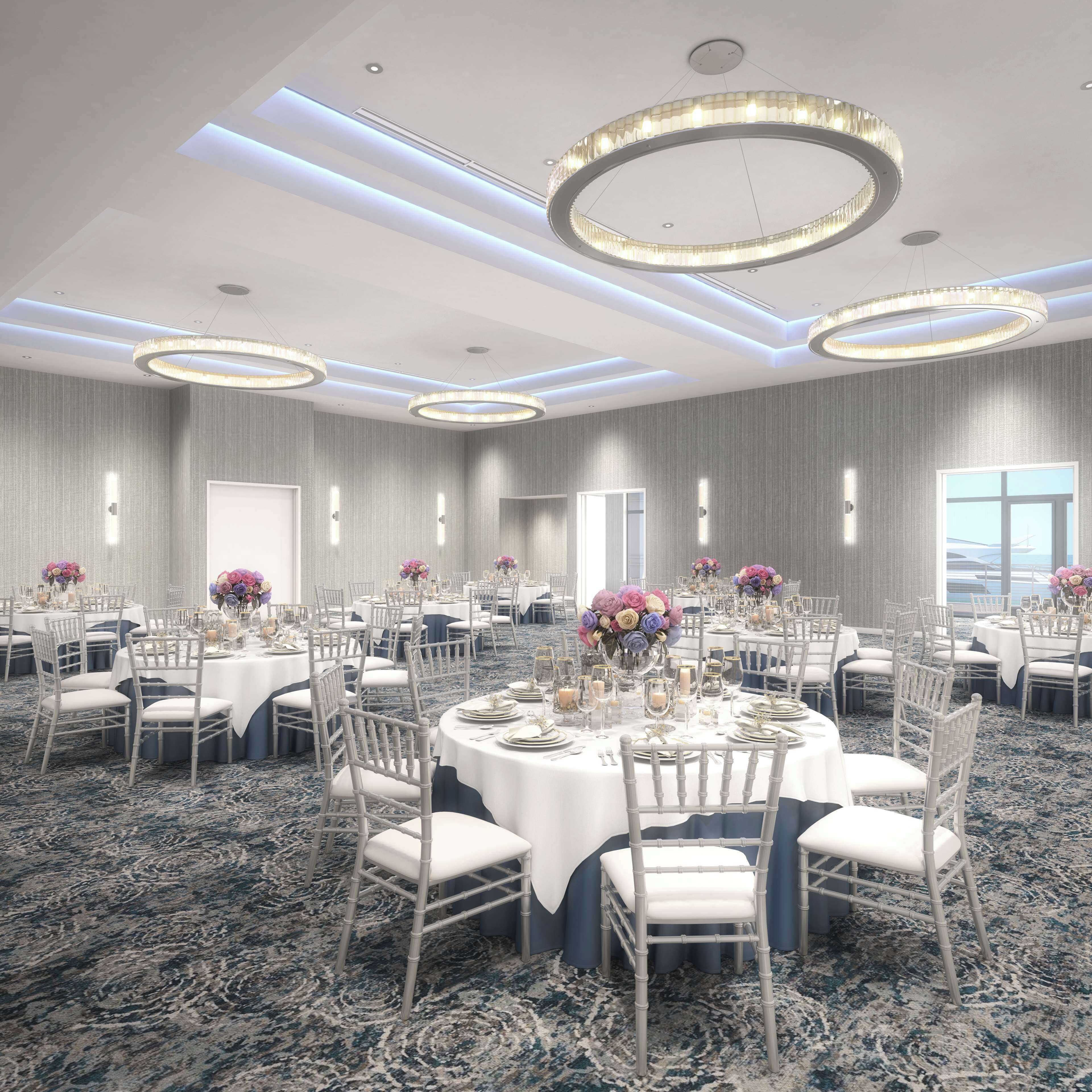 A spacious banquet hall with round tables, white chairs, and chandeliers.