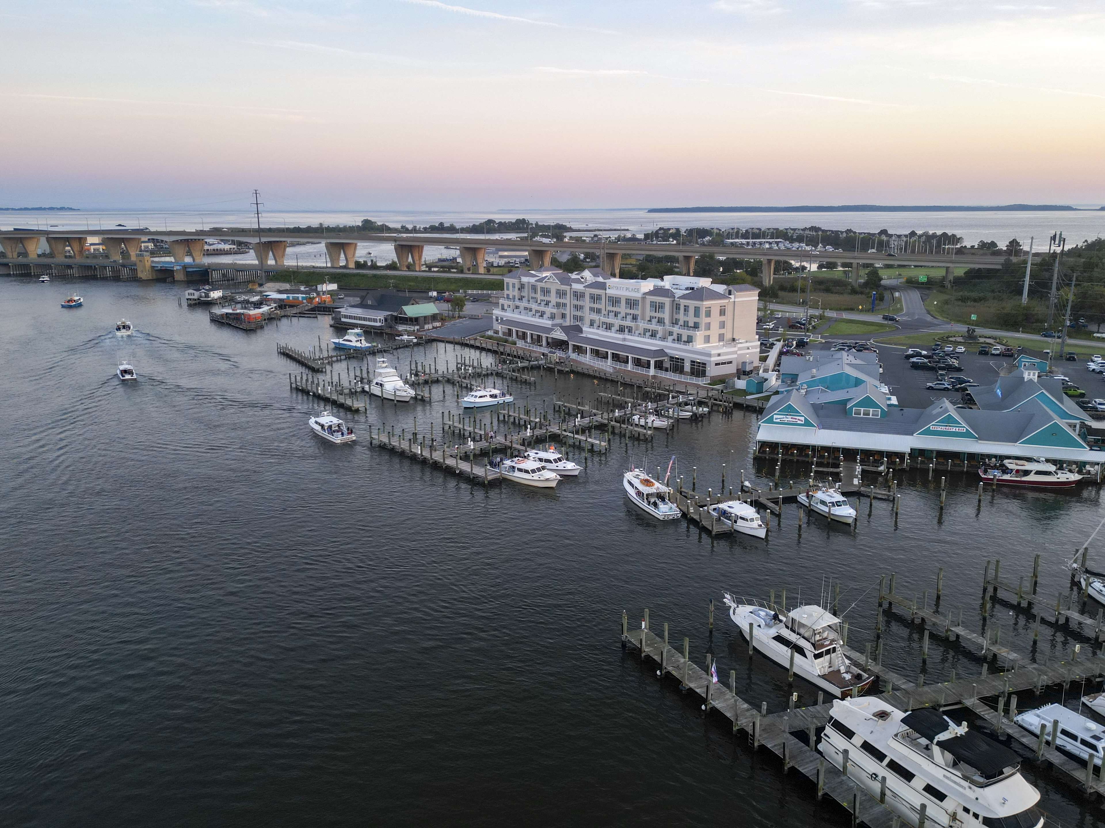 A marina with multiple docks and boats, a large building with a sign reading "Marriott", and a bridge in the background.