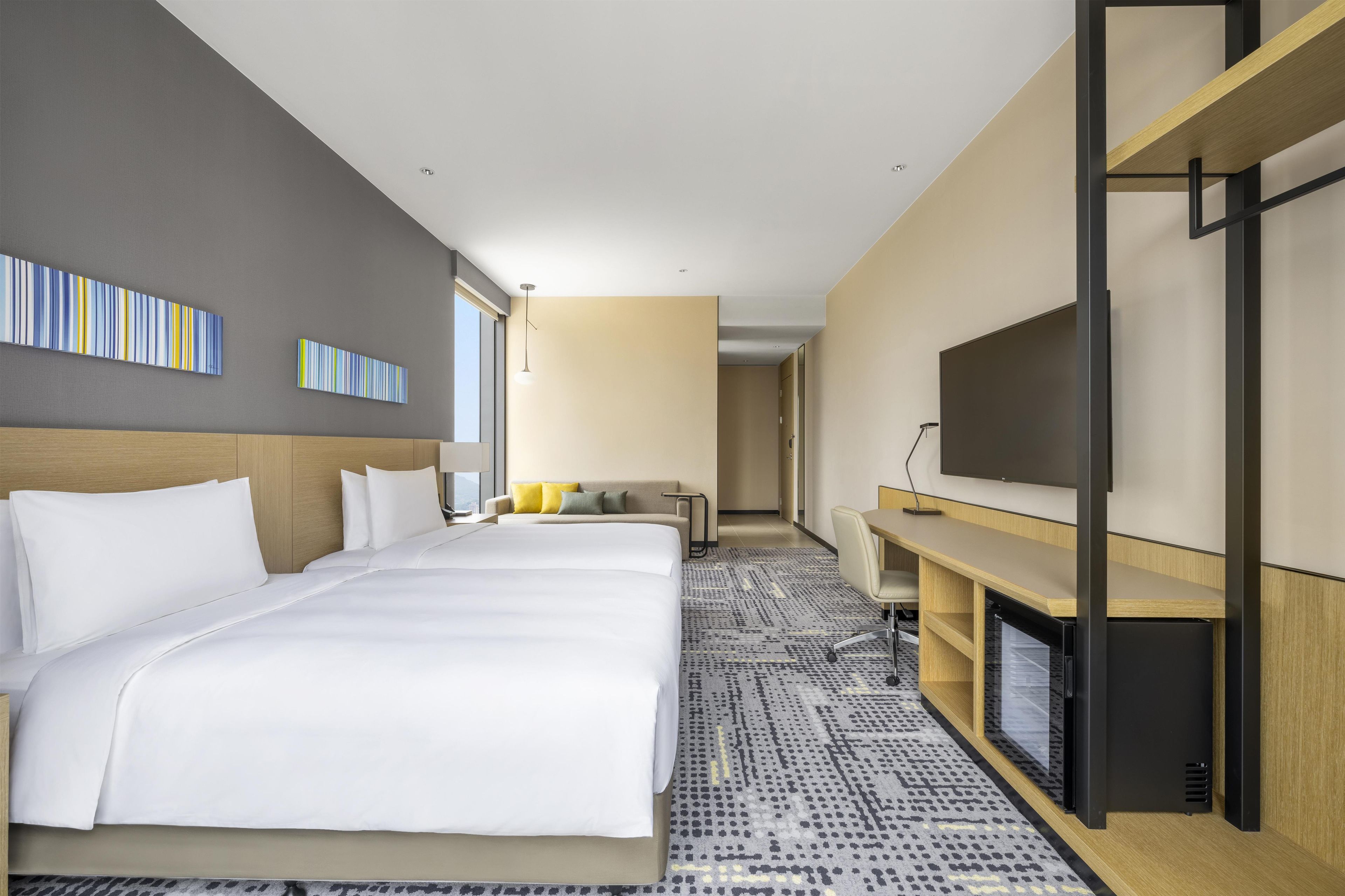 A hotel room with two beds, a TV, a desk, and a large window.
