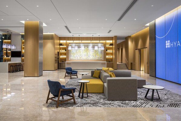 Un hall d’hôtel moderne, doté d’espaces aménagés avec des sièges, d’un comptoir d’accueil et d’un mur bleu sur lequel figure le nom de l’hôtel.
