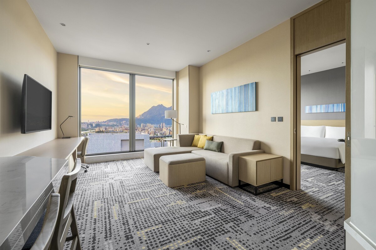 Une chambre d’hôtel moderne dotée d’une grande fenêtre donnant sur la ville, d’un téléviseur à écran plat, d’un canapé, d’un bureau et d’un lit.