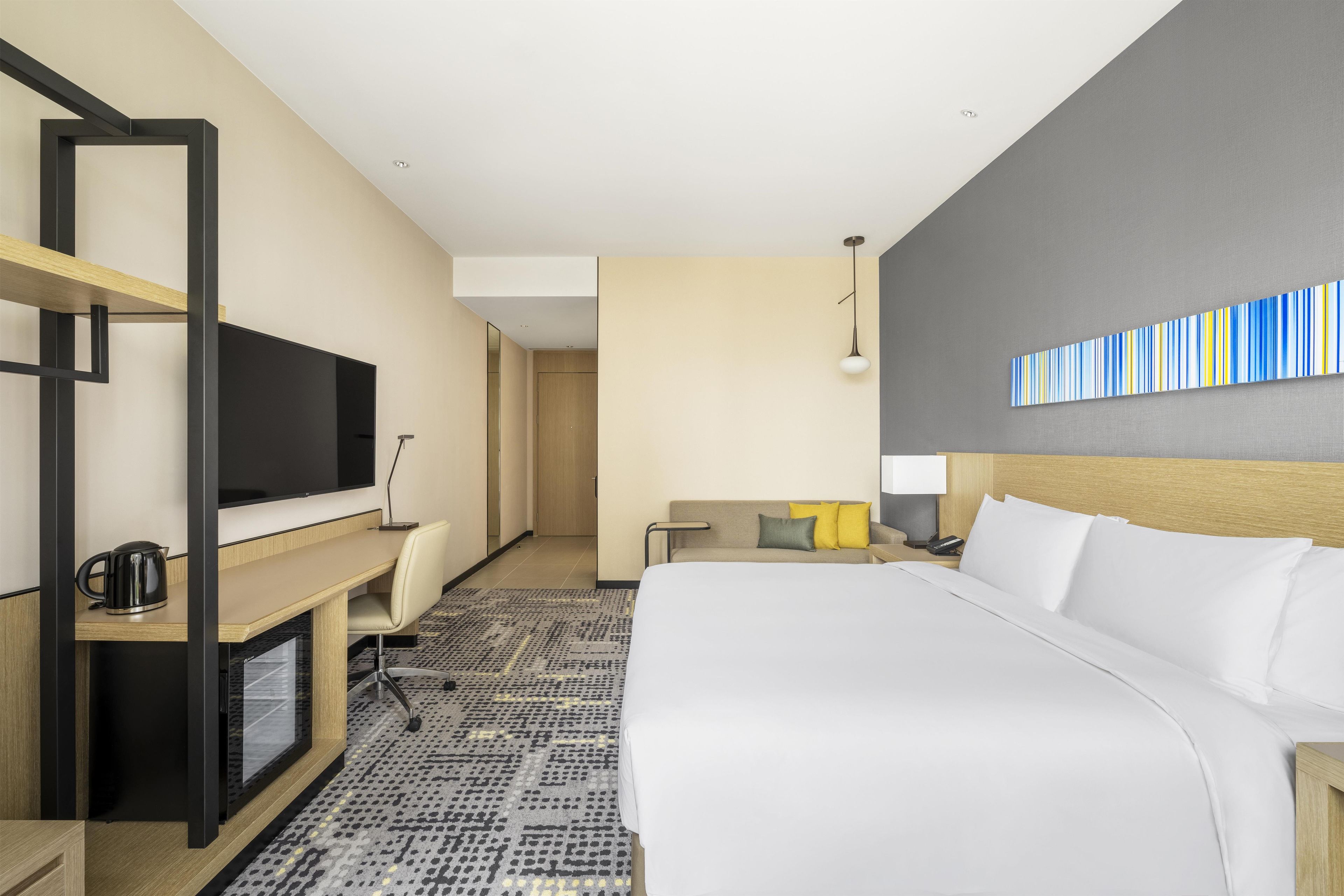 A hotel room with a bed, a desk, a television, a sofa, a patterned rug, and a wall art piece.